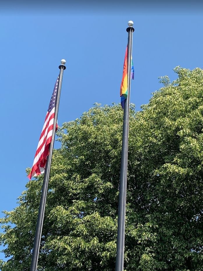 Pride flag and American flag side by side at the top of flagpoles in front of the municipal bluiding